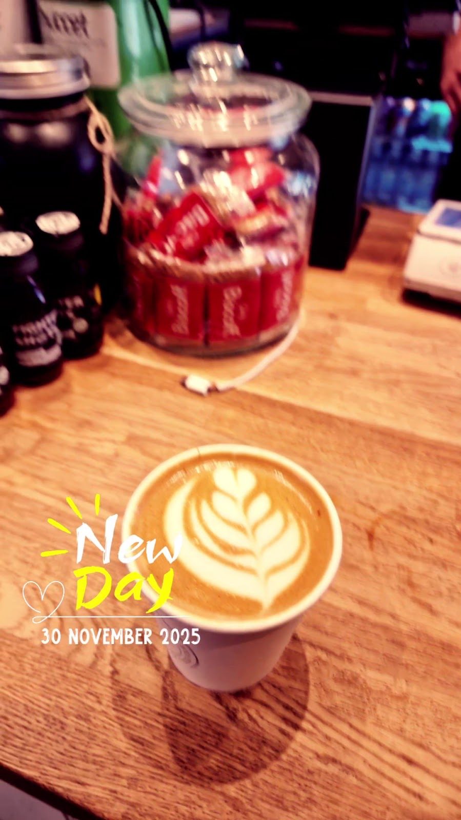 A takeaway latte with leaf art on the counter, sweet jars behind